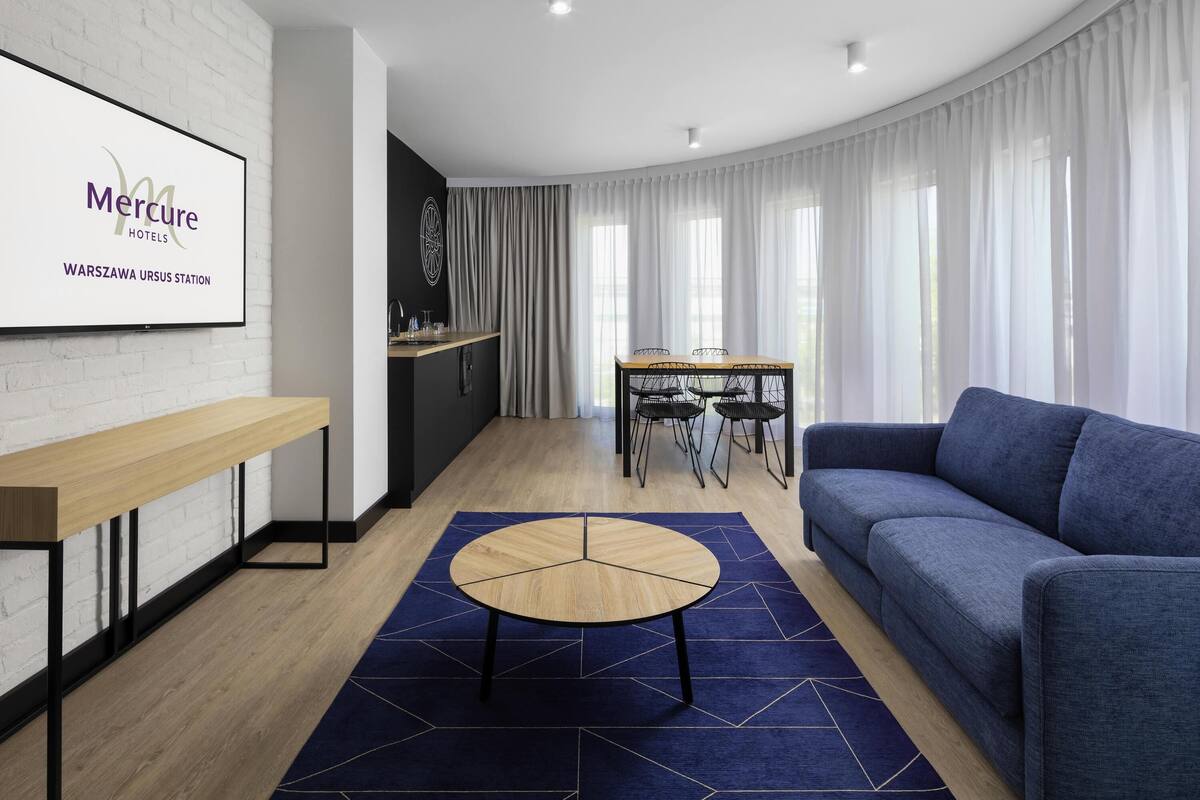 Une chambre d’hôtel moderne dotée d’un coin repas, d’un canapé et d’un téléviseur affichant le nom de l’hôtel.
