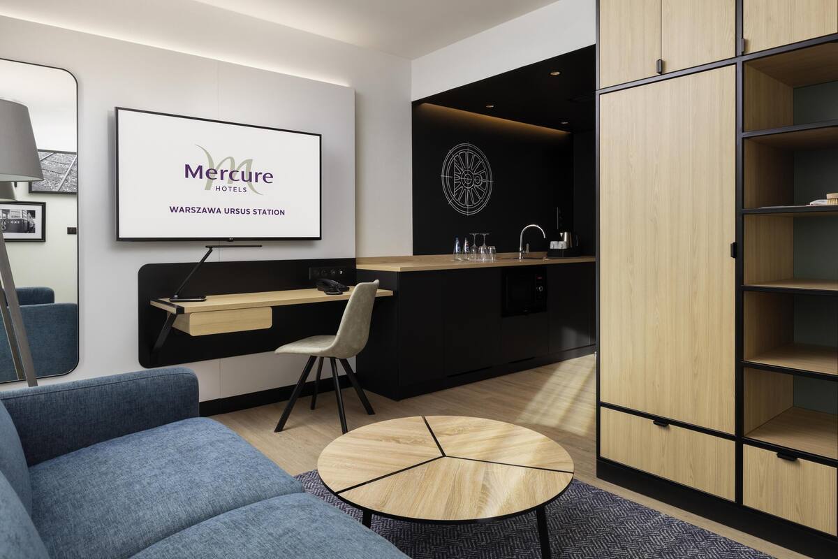 Une chambre d’hôtel moderne comprenant un canapé, une table ronde, un bureau avec une chaise, un téléviseur fixé au mur, un coin cuisine avec un évier et une armoire.