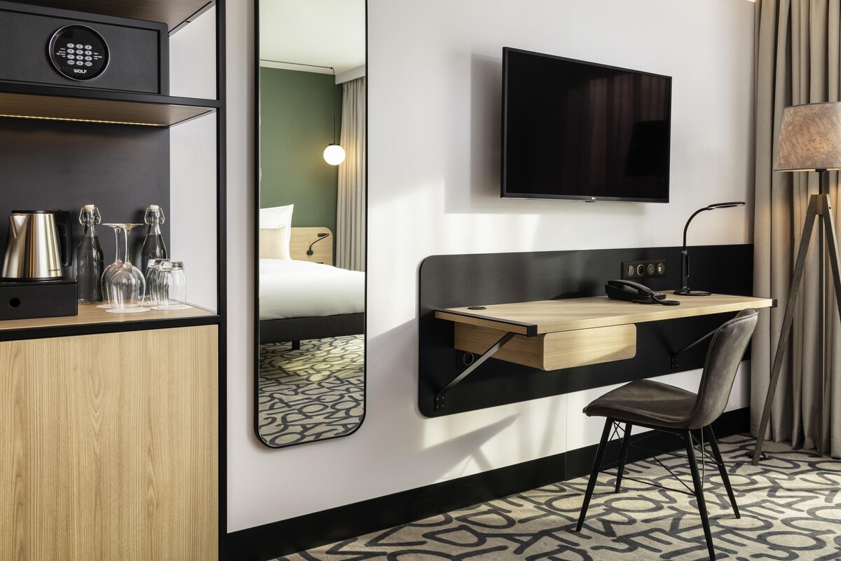 A hotel room with a desk, chair, TV, mirror, and a coffee maker on the side table.