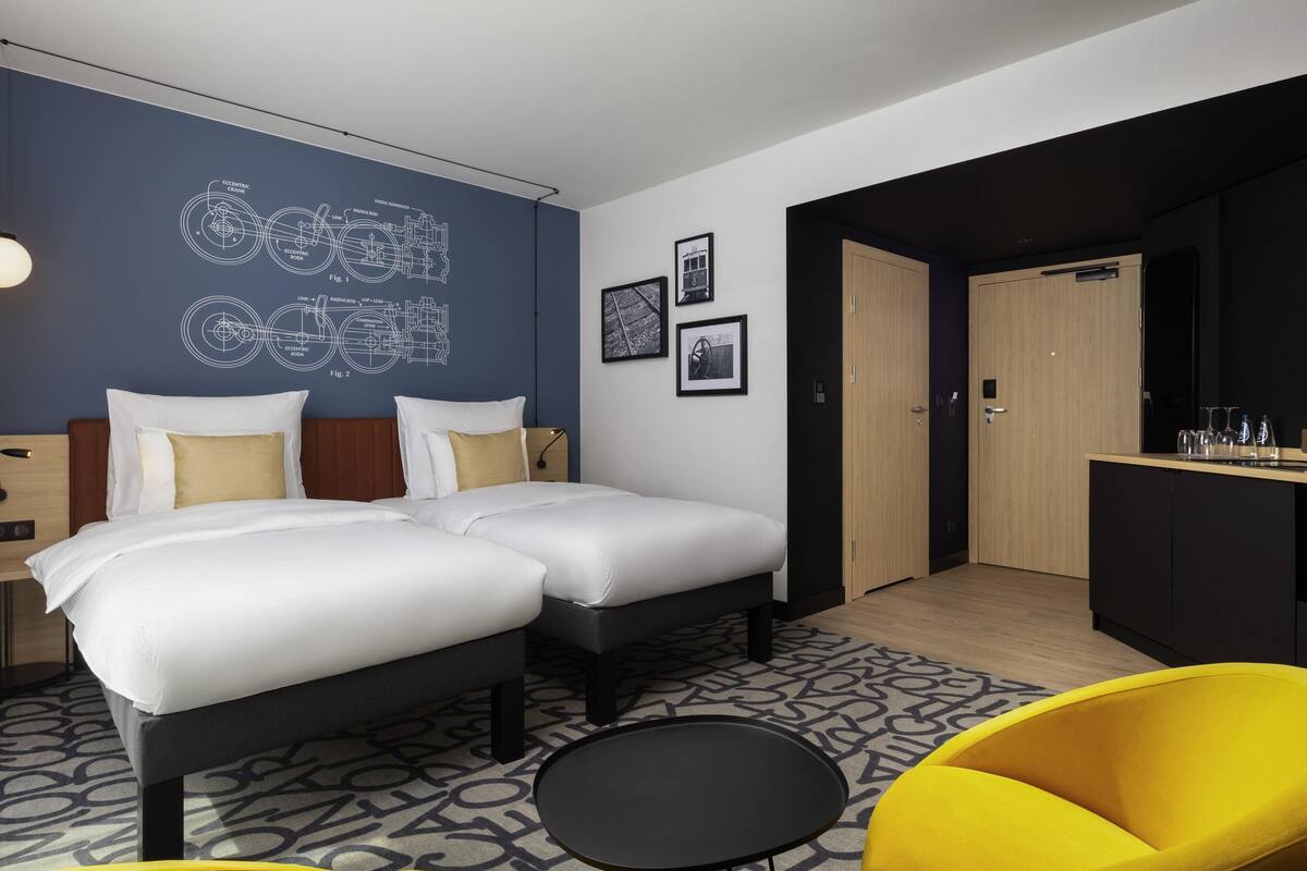Une chambre d’hôtel moderne avec deux lits, un petit coin repas et un espace salon avec des chaises jaunes.