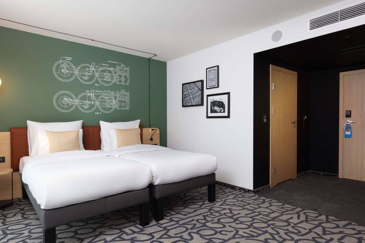 Une chambre d’hôtel avec un lit, un mur vert orné de schémas techniques, des tableaux encadrés et une porte.