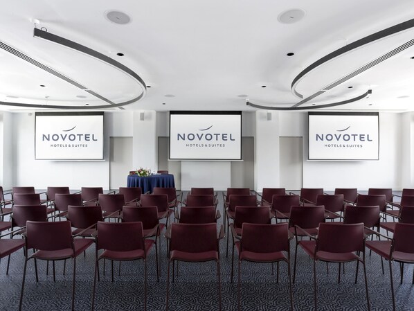 A conference room with rows of chairs, a table with a blue tablecloth, and three screens displaying the Novotel logo. - Novotel Convention And Spa (Antananarivo)