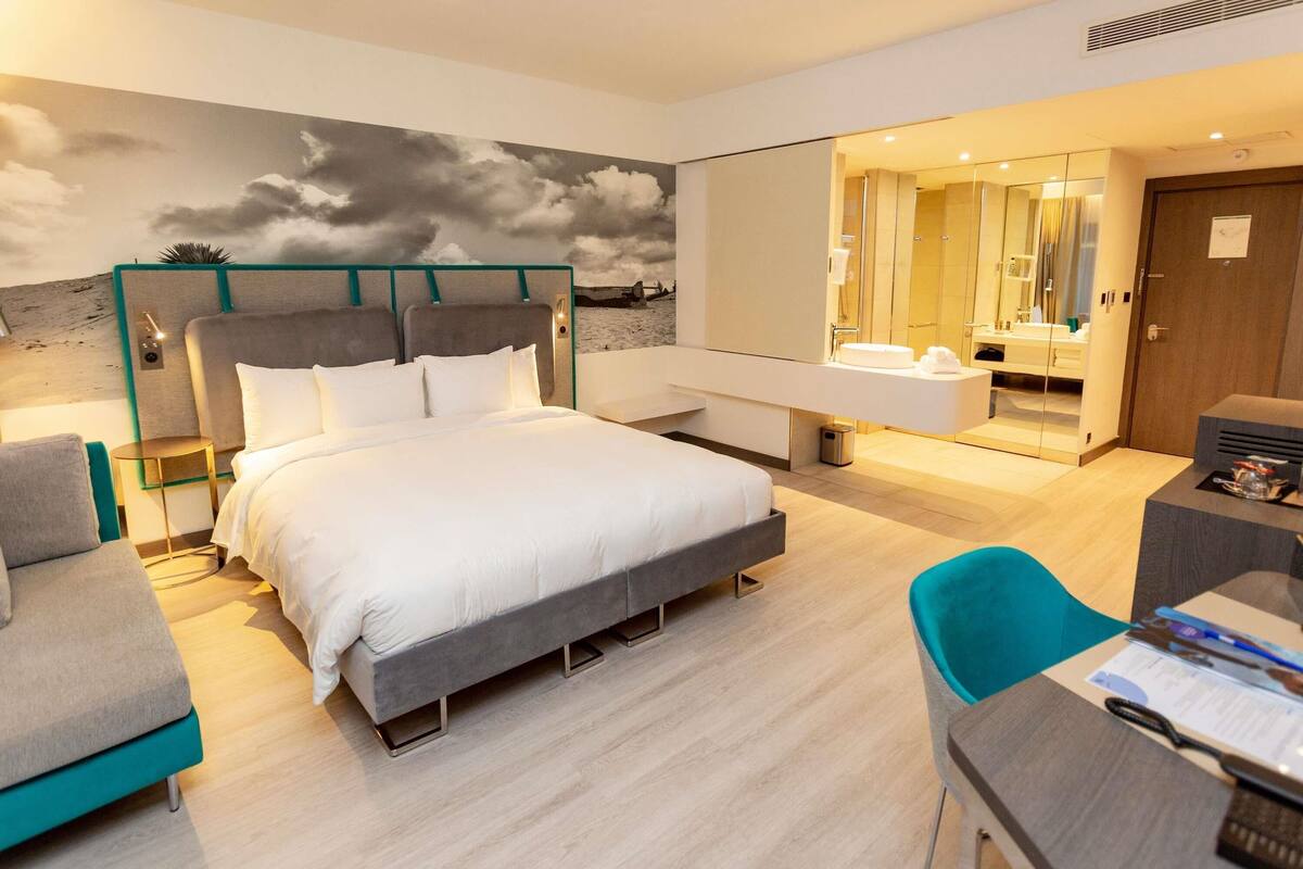 A modern hotel room with a bed, a desk, a chair, a bathroom with a mirror, and a wall mural.