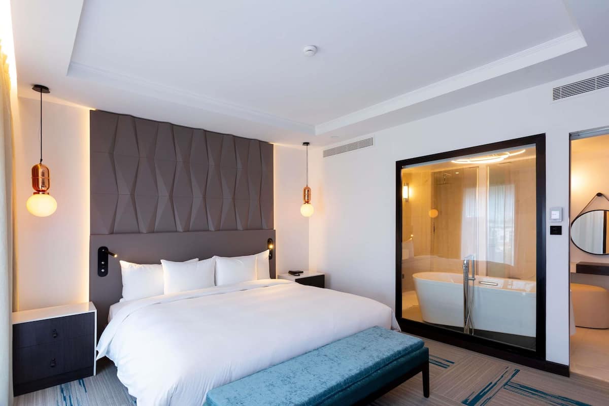 Ein modernes Hotelzimmer mit einem großen Bett, einer Sitzbank, einer Duschkabine aus Glas und einem kleinen Nachttisch.