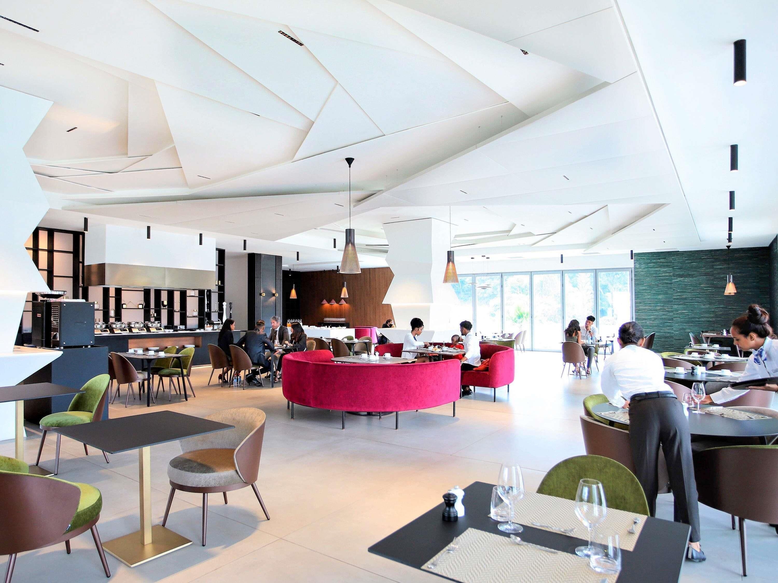 A modern dining area with tables and chairs, a central red seating area, and a kitchen in the background.