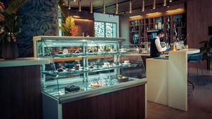 A dessert display case in a modern cafe with a wooden counter and a person working behind it. - Novotel Convention And Spa (Antananarivo)