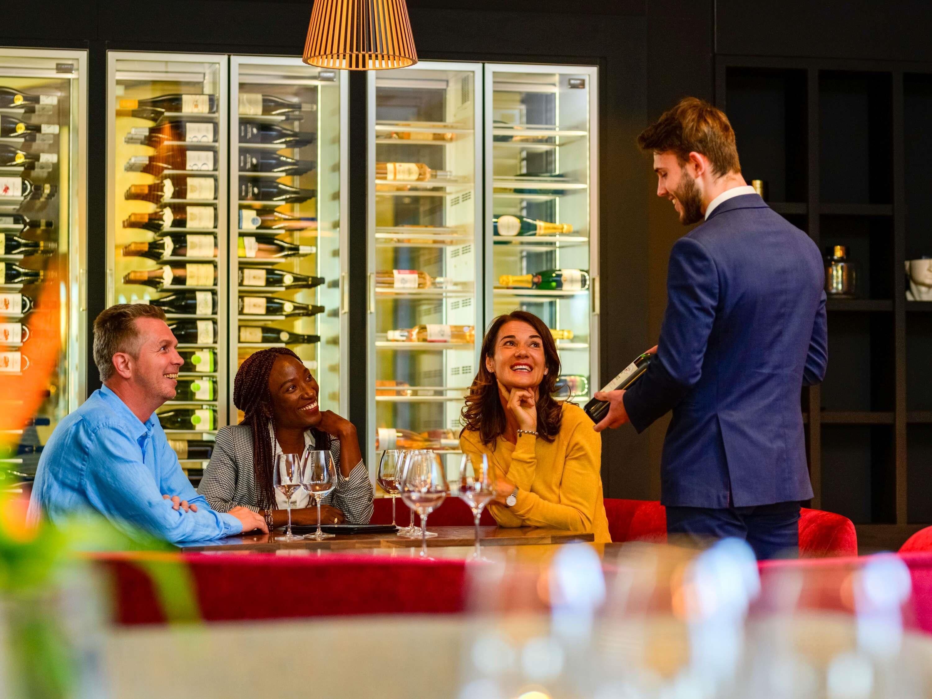 A group of people seated at a table with wine glasses, a man standing with a bottle, and a wine refrigerator in the background.