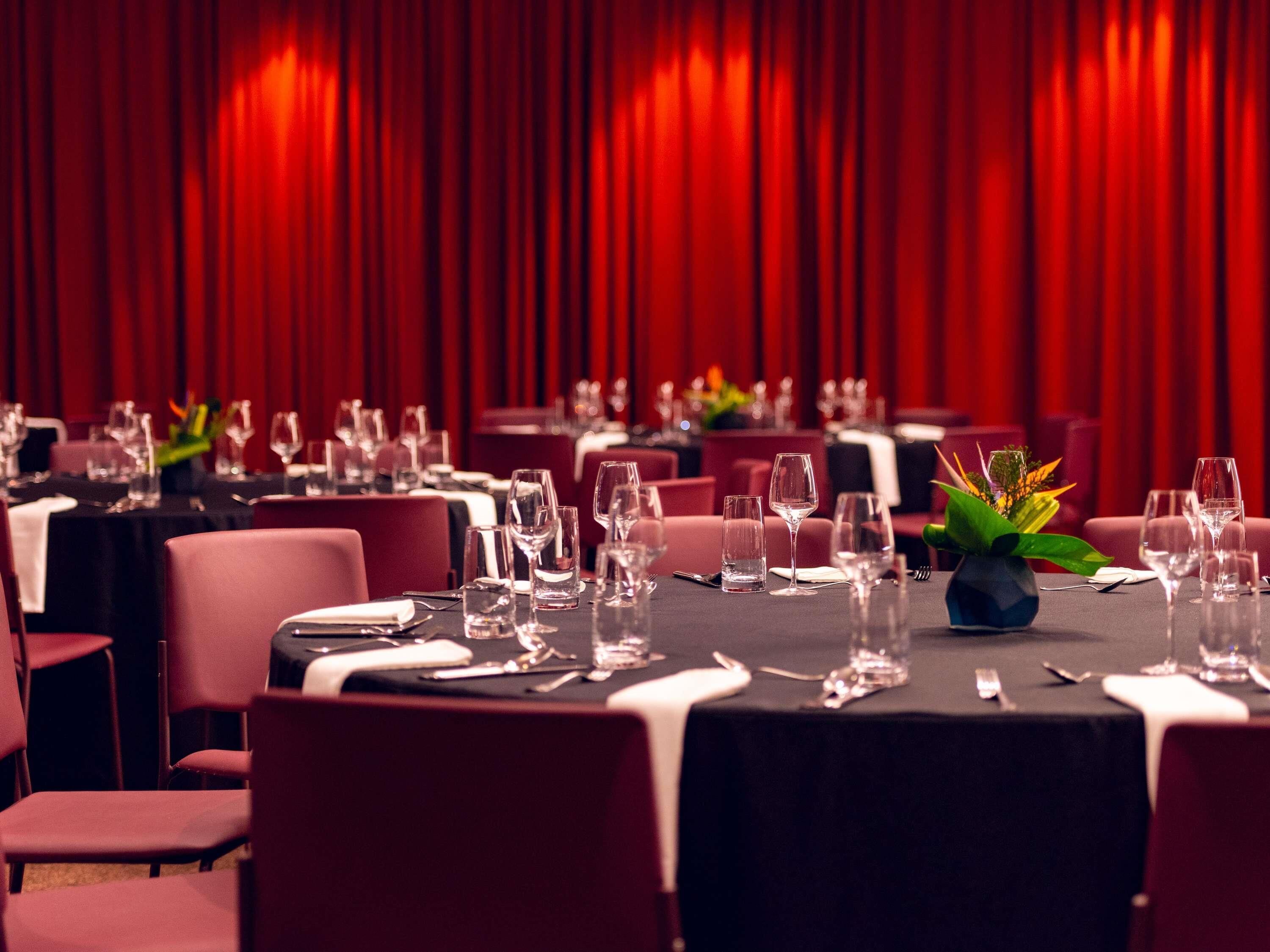 Een restaurant met gedekte tafels, rode gordijnen op de achtergrond en een vaas met een plant op een tafel.