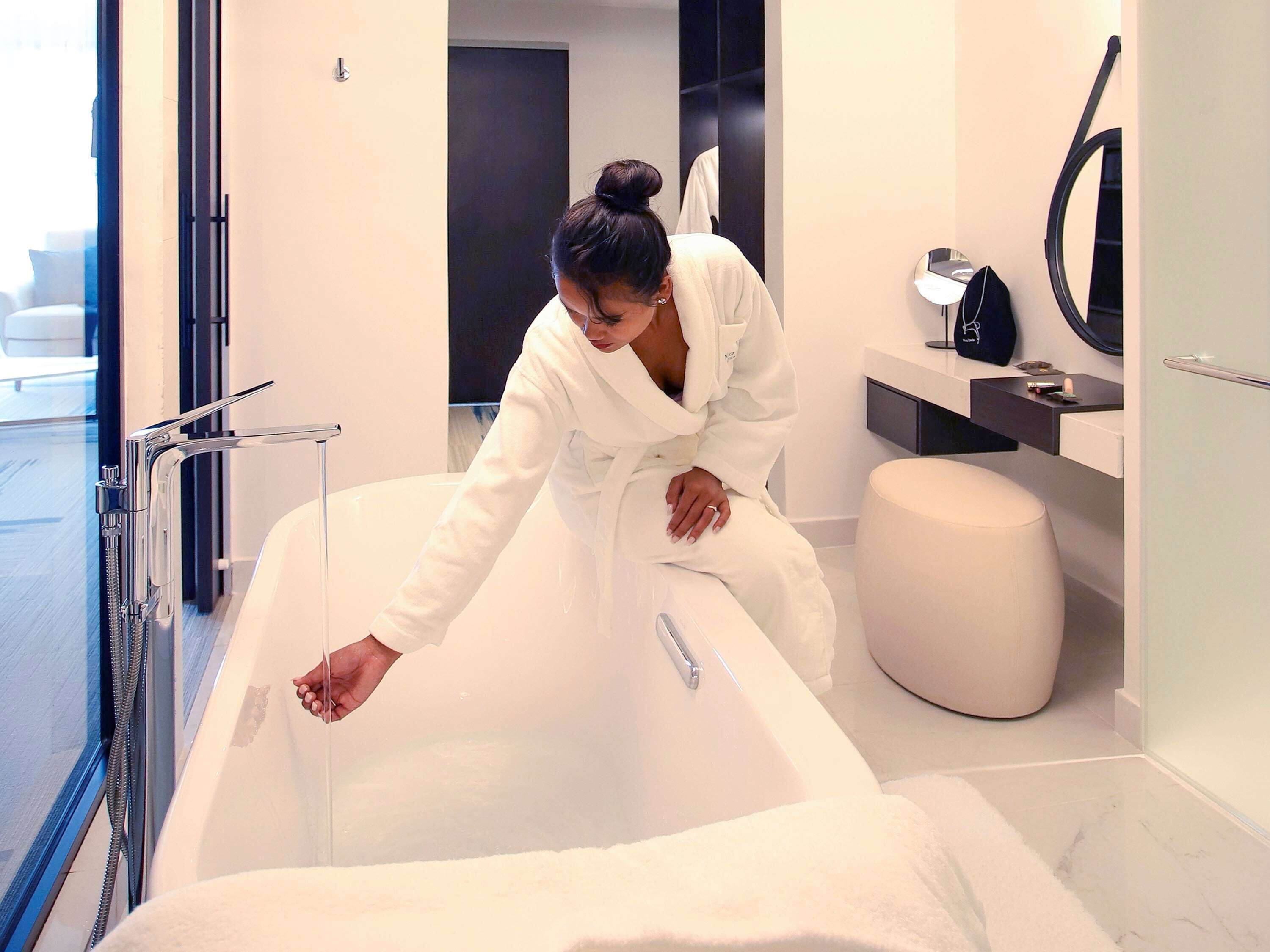 A person in a white bathrobe leaning over a bathtub in a modern bathroom with a mirror, a towel, and a small shelf with toiletries.