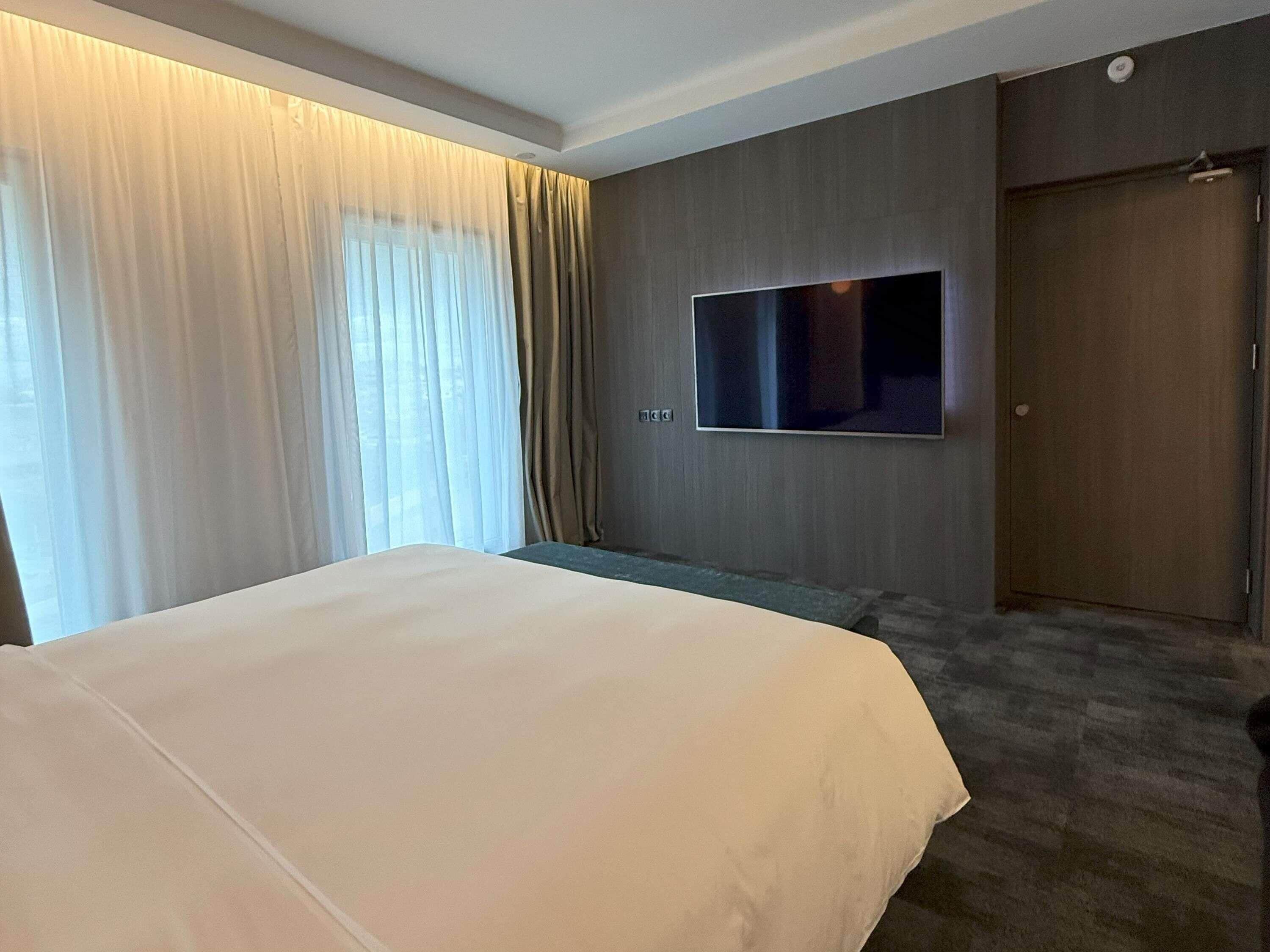 A hotel room with a large bed, a flat-screen TV mounted on the wall, a door, and a window with curtains.