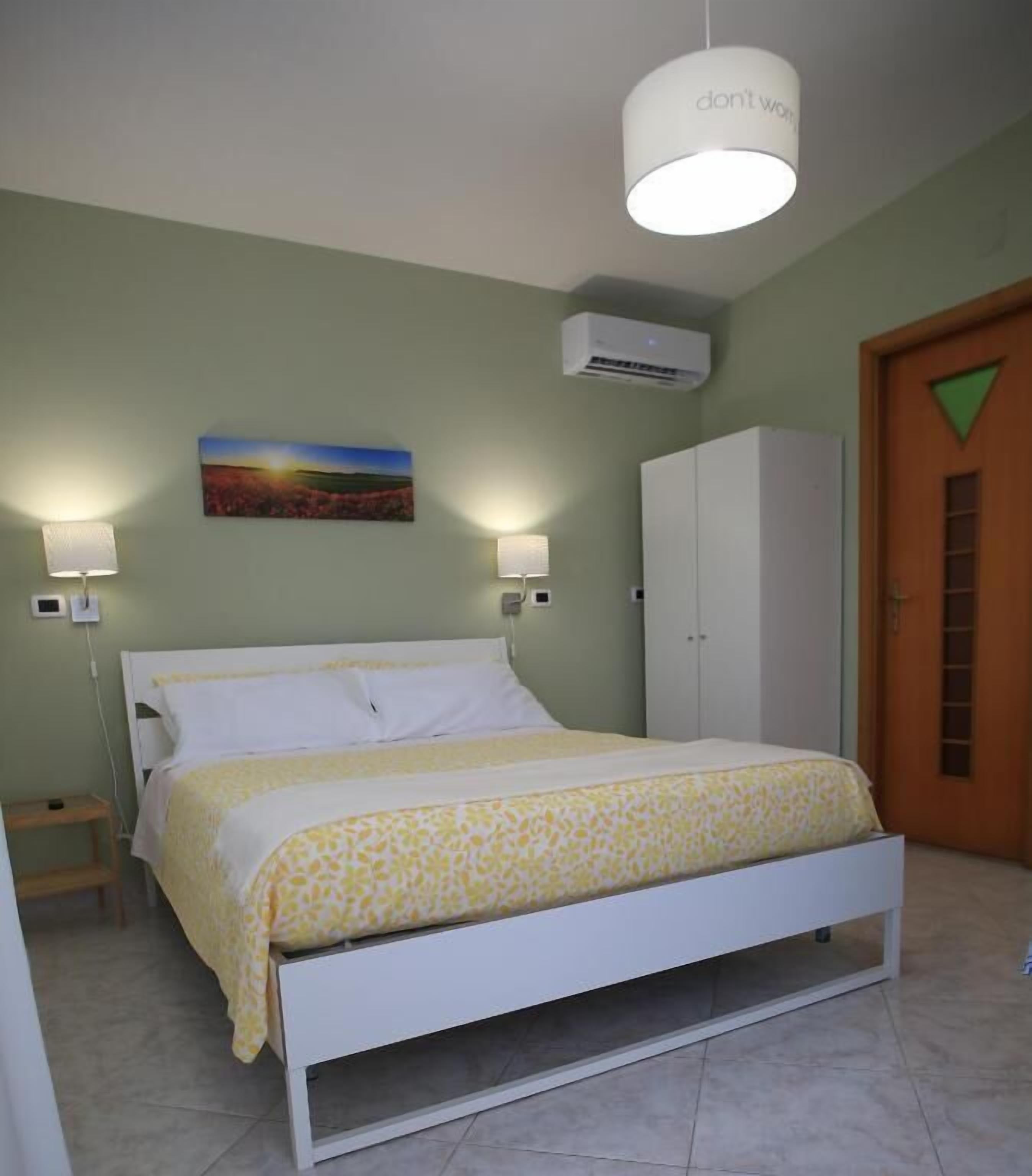 A bedroom with a bed, two bedside lamps, an air conditioning unit, a wardrobe, and a door with a green triangle.