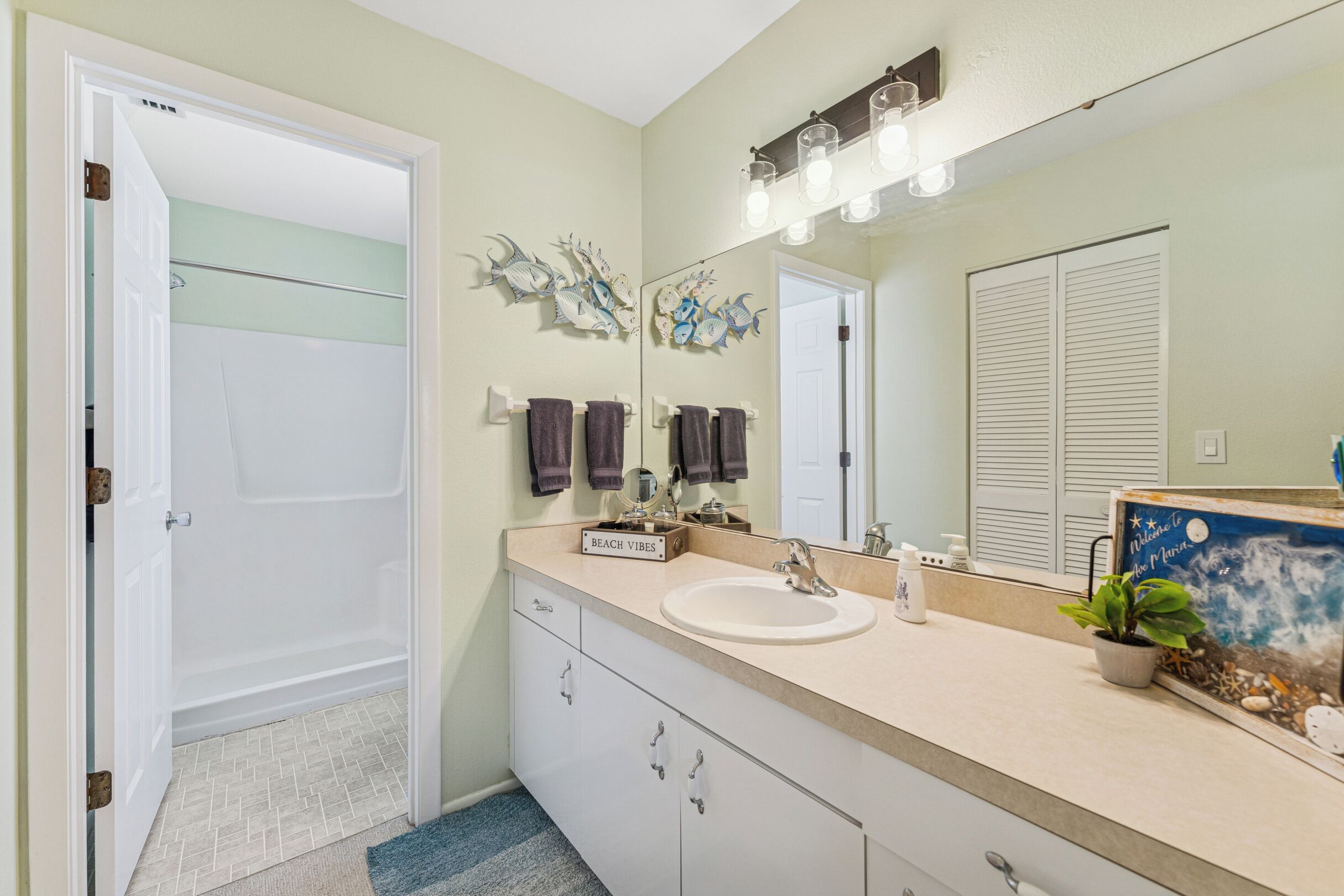 A bathroom with a white sink, a mirror, and a shower. There are towels hanging on the wall and a small plant on the counter.