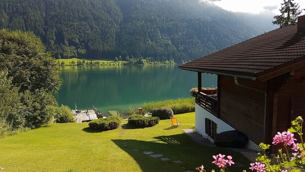 A lakeside cabin with a wooden deck, a small boat, and a clear view of a lake surrounded by forested mountains. - Chalets Zöhrer - Wohnen am Wasser (Weissensee)