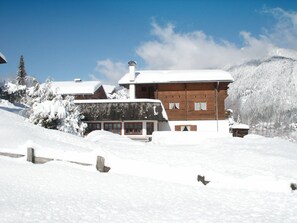A wooden chalet with a snow-covered roof, surrounded by a snowy landscape and mountains.