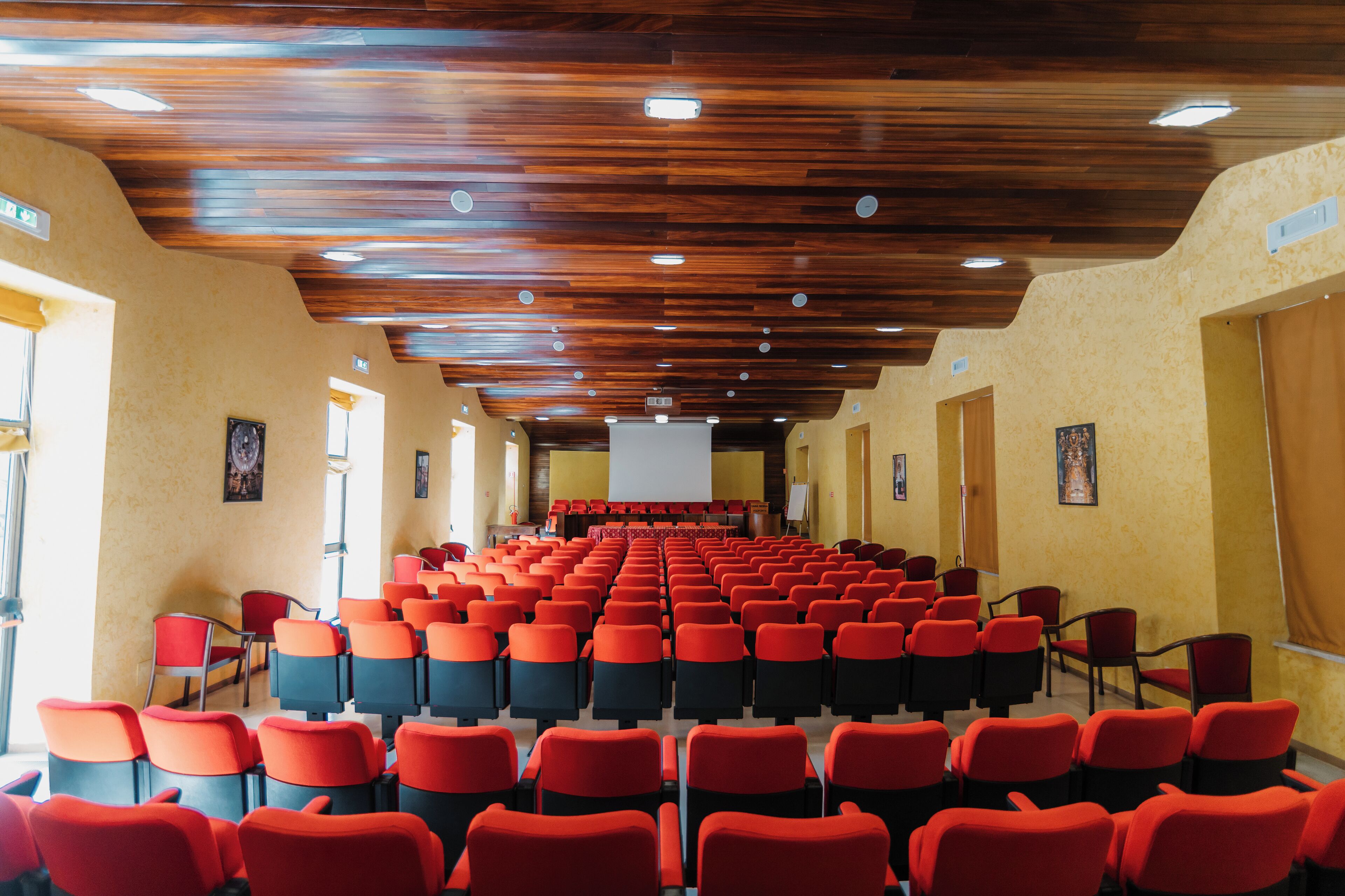 A conference room with rows of red chairs, wooden ceiling, and beige walls.