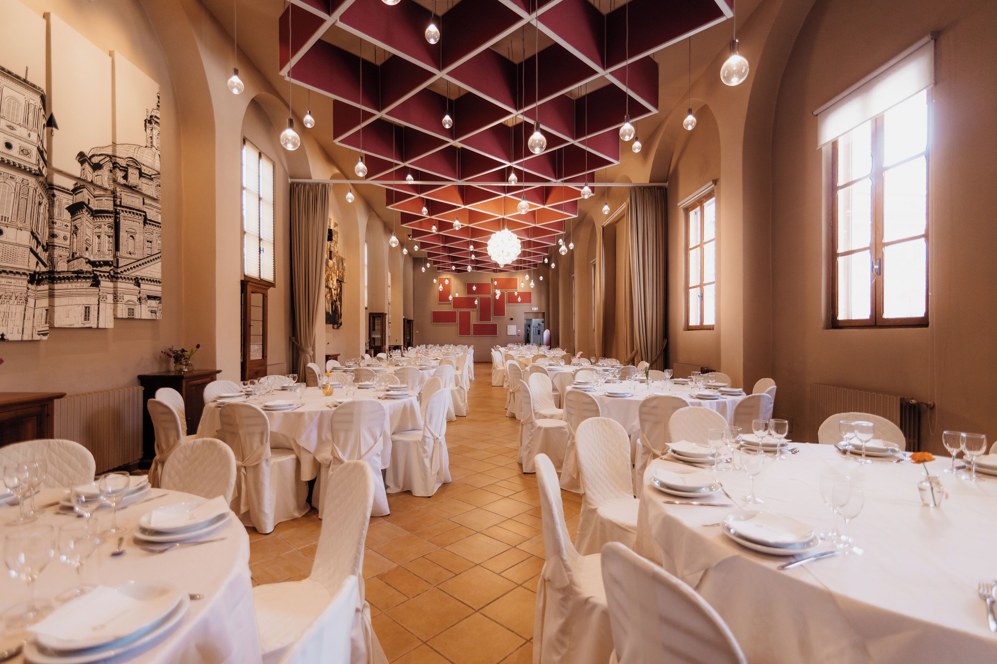 A banquet hall with round tables set for dining, large windows, and a high ceiling with hanging lights.
