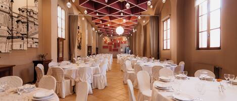 A banquet hall with round tables set for dining, large windows, and a high ceiling with hanging lights.