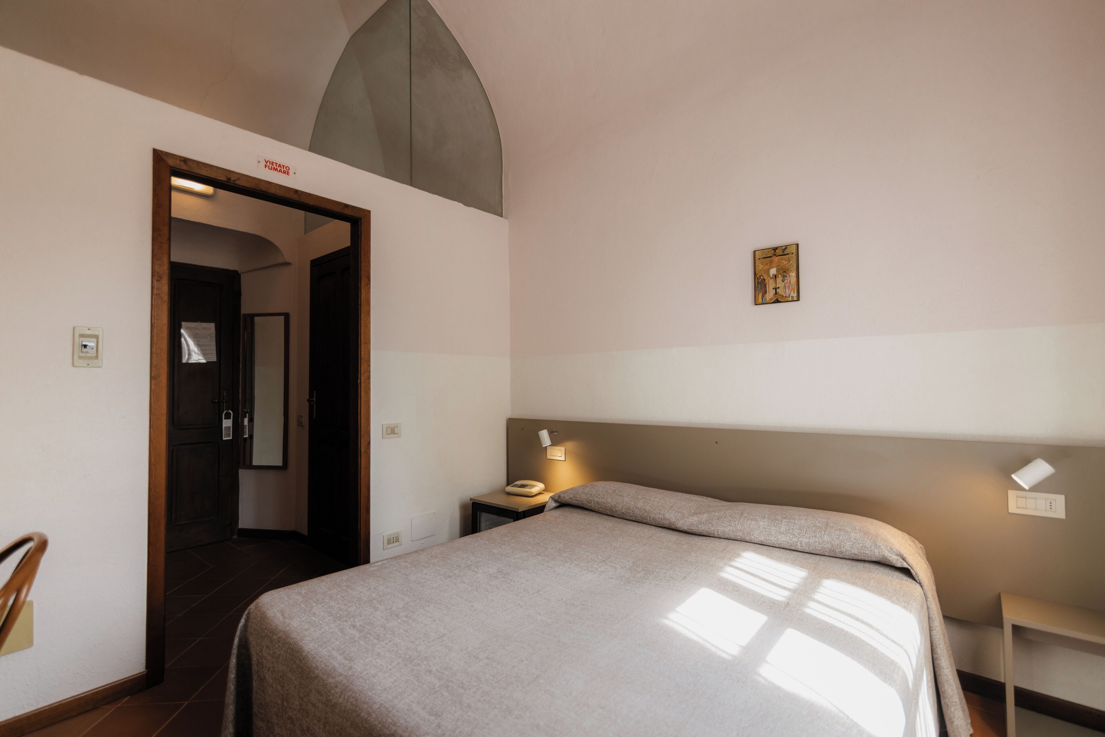 Habitación de hotel con cama, mesita de noche, silla, puerta y un cuadro en la pared.