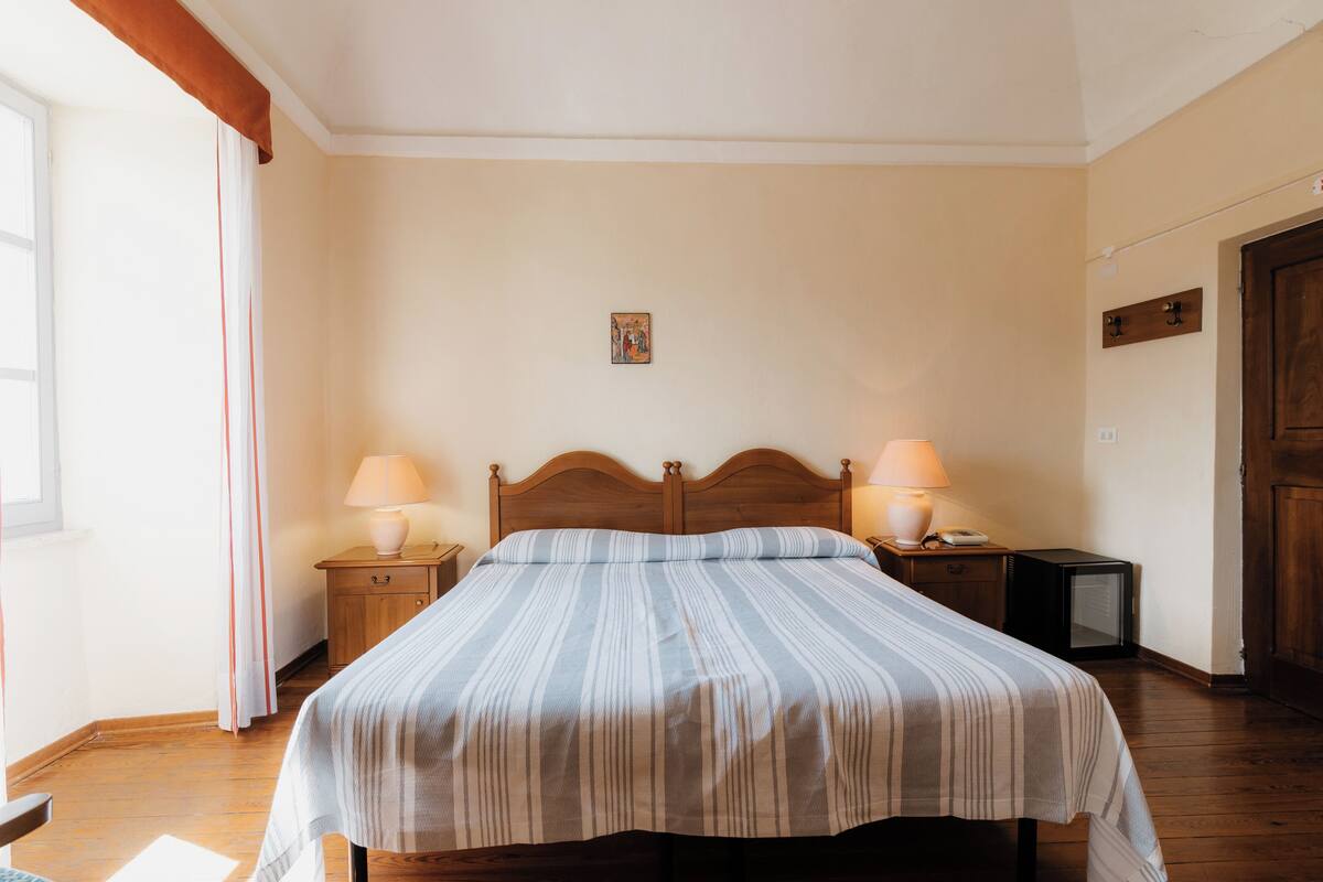 A bedroom with a wooden bed, two bedside tables with lamps, a small bedside table with a phone, a wooden wardrobe, and a window with curtains.