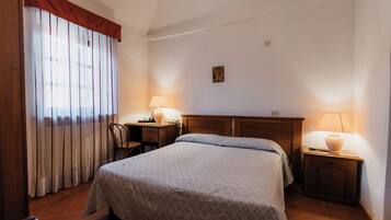 A bedroom with a bed, two bedside tables with lamps, a desk with a chair, and a window with curtains.