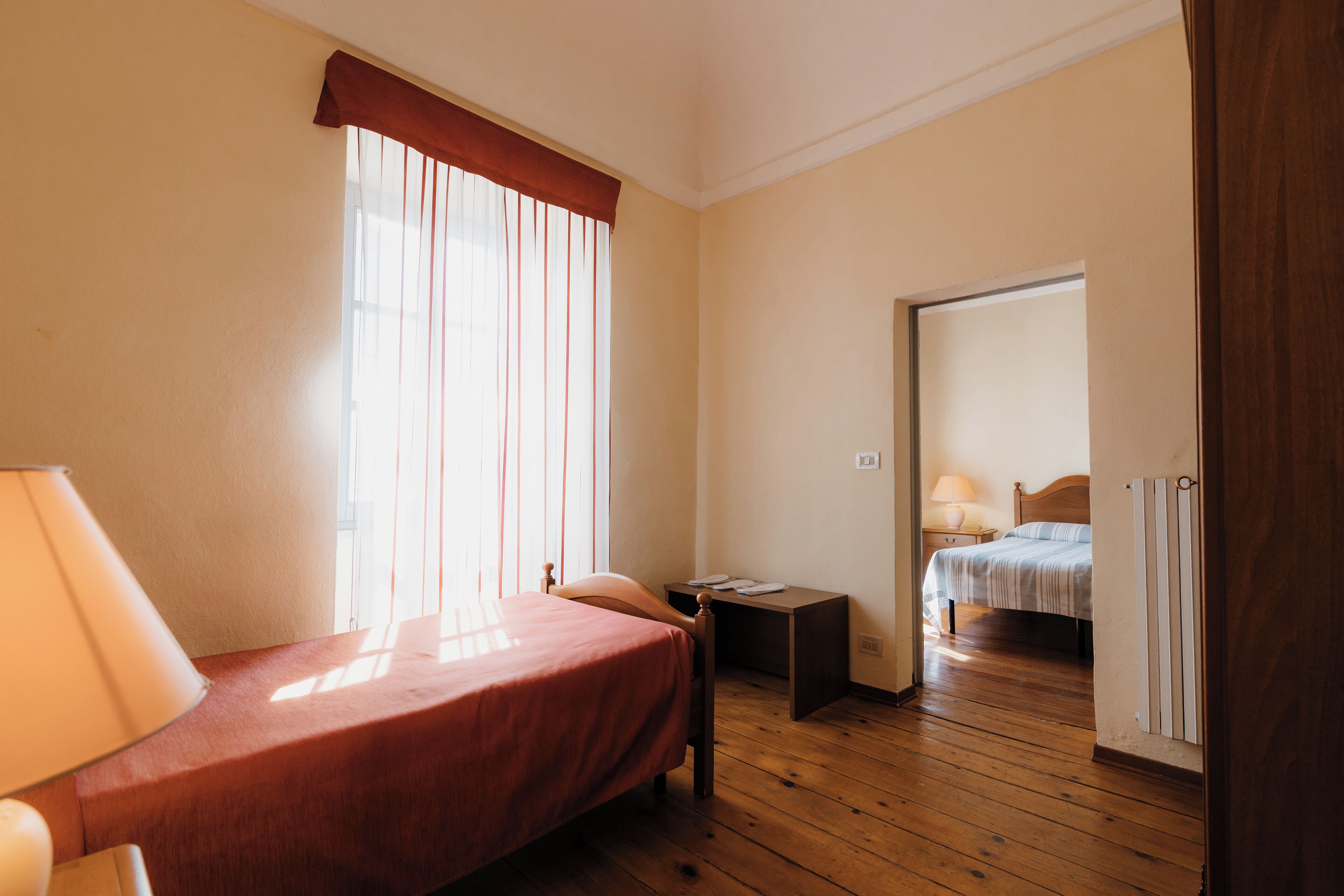 Un dormitorio con una cama, una mesita de noche, una lámpara, una ventana con cortinas y suelo de madera.
