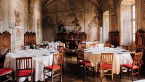 Un vaste réfectoire orné de fresques au plafond, de boiseries et d’un autel central. Les tables sont dressées avec des nappes blanches et des chaises rouges.