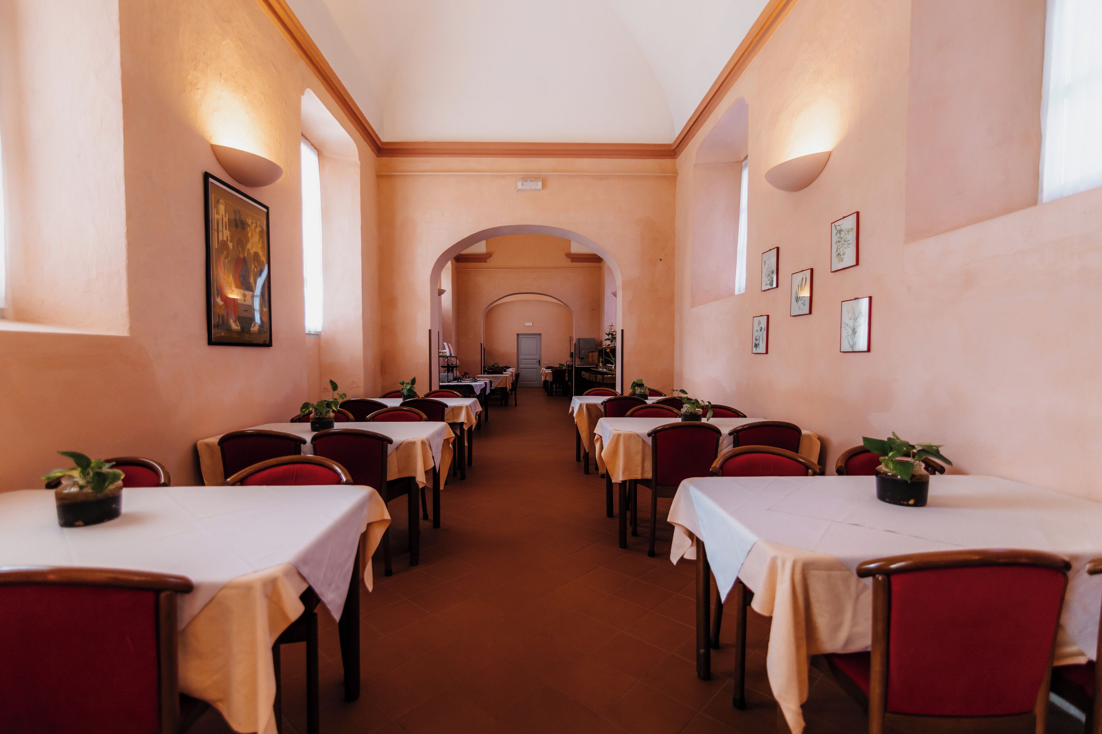 A hotel dining area with tables and chairs arranged in rows, a central archway, and framed pictures on the walls.