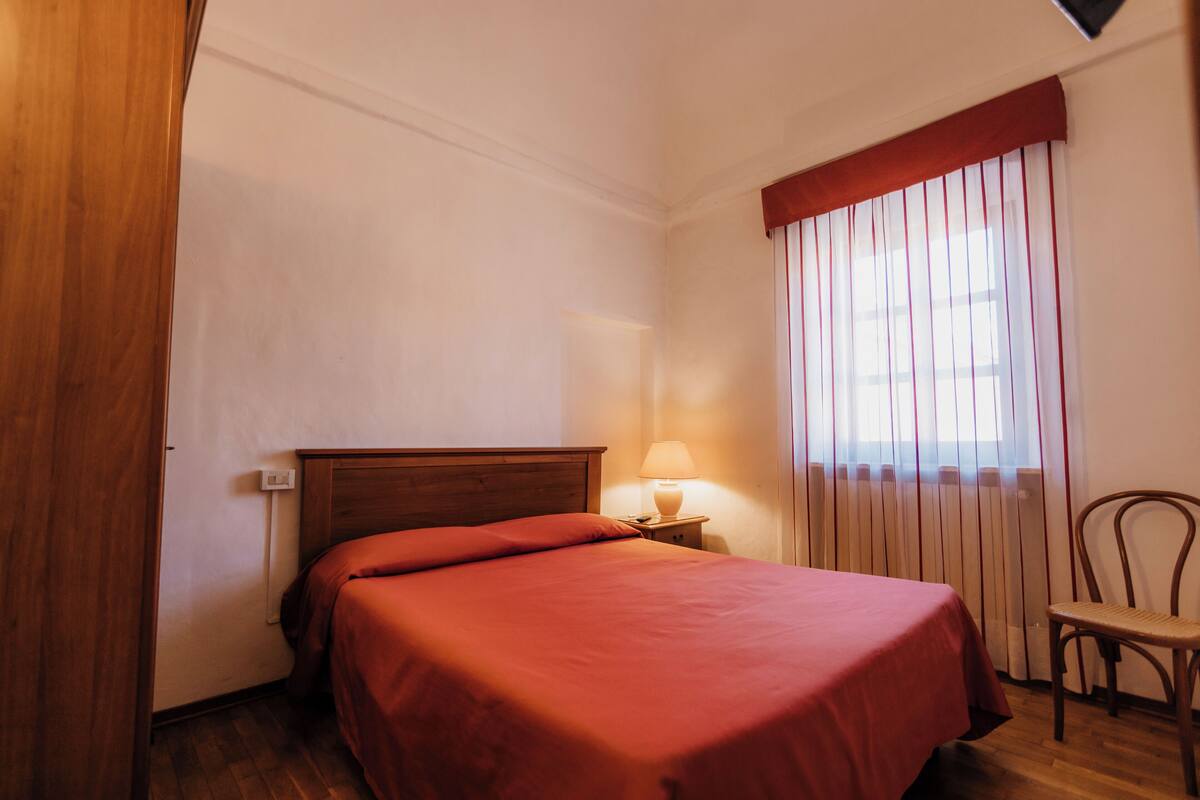 A bedroom with a wooden bed, a nightstand with a lamp, a chair, and a window with vertical blinds.