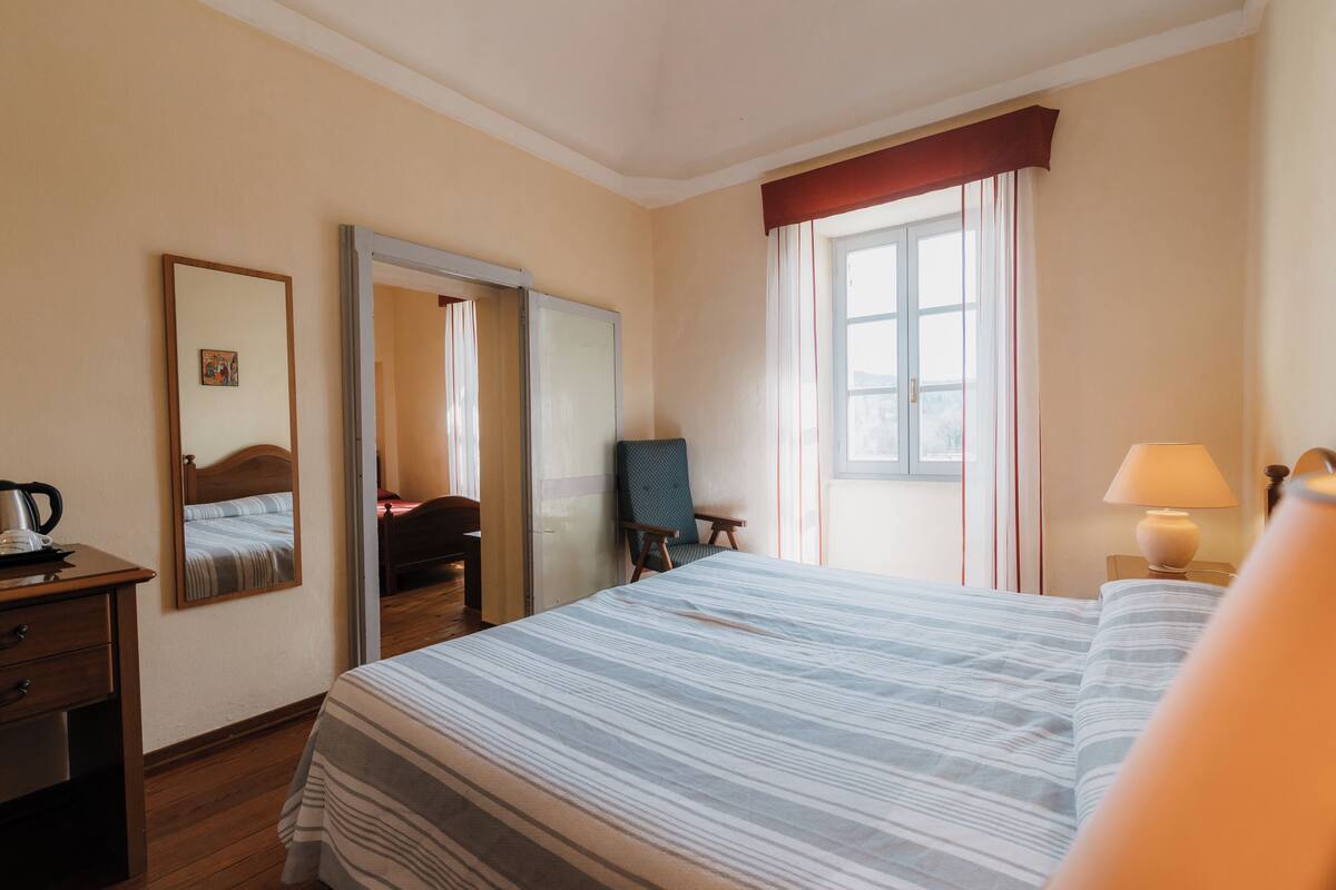 A hotel room with a bed, a desk with a lamp, a chair, a mirror, and a window with curtains.