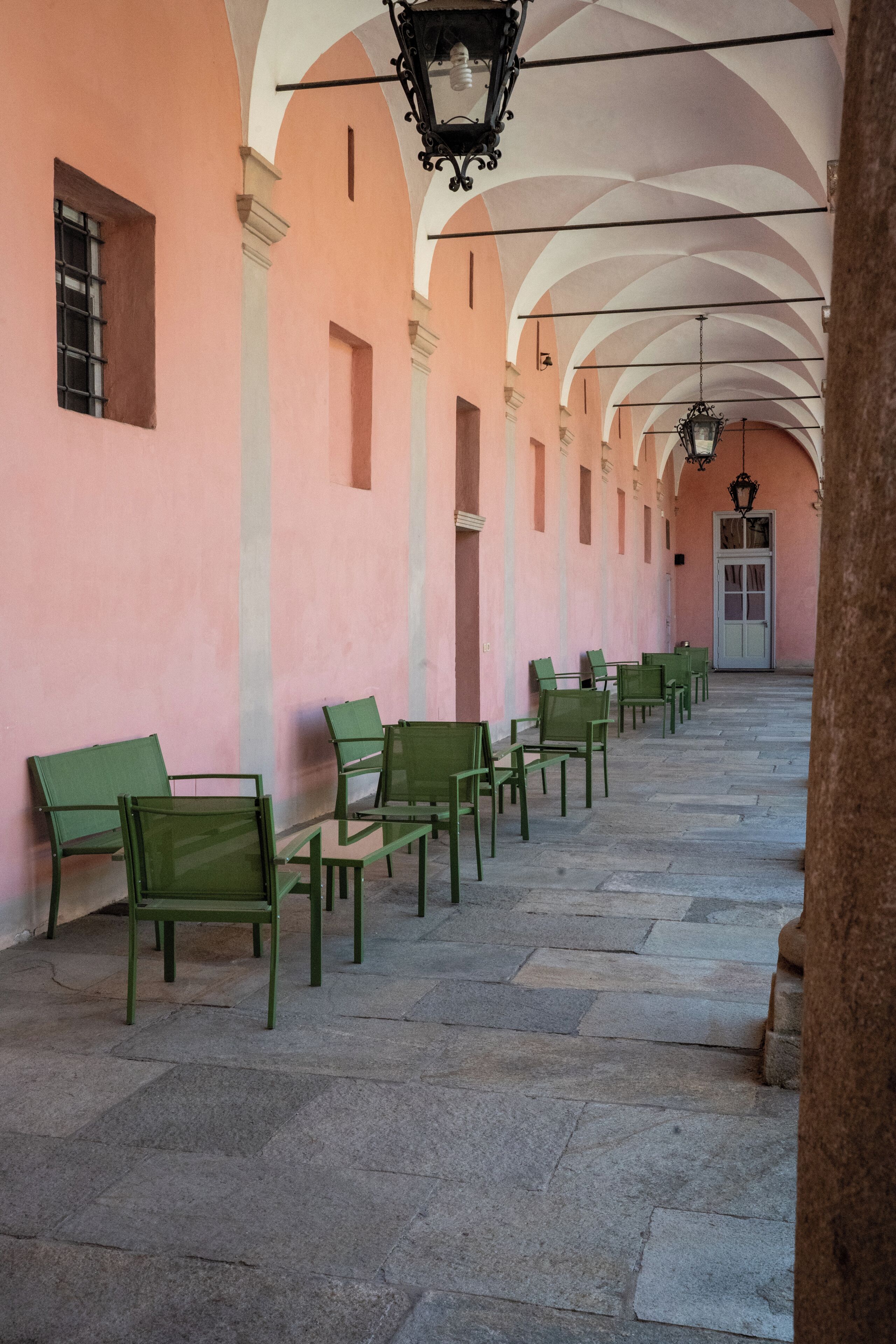 Un corridoio con sedie verdi disposte lungo le pareti, un pavimento in pietra e una serie di archi con lanterne sospese.