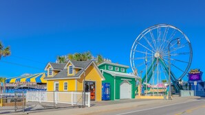 Un parque de atracciones en la costa con una noria grande, un edificio amarillo con el rótulo 'Hurricane' y un edificio verde con un cartel de Pepsi.