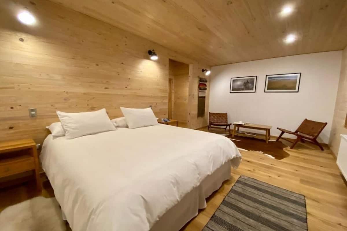 Un dormitorio con paneles de madera, una cama grande, dos sillas, una mesita y una alfombra.