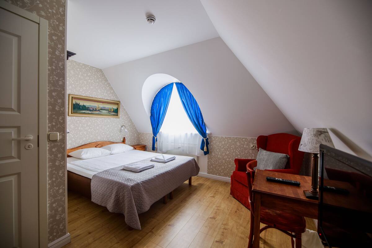 A bedroom with a bed, a desk, a chair, a lamp, and a window with blue curtains.