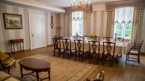 A dining room with a wooden table set for six, surrounded by six chairs. There is a chandelier above the table, and a large window with curtains on the right.