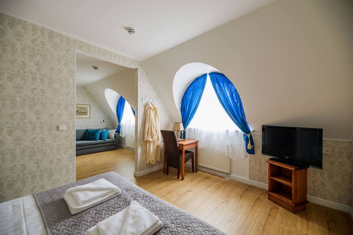 A bedroom with a bed, a TV on a wooden stand, a desk with a chair, and a view of a living area through a window with blue curtains.