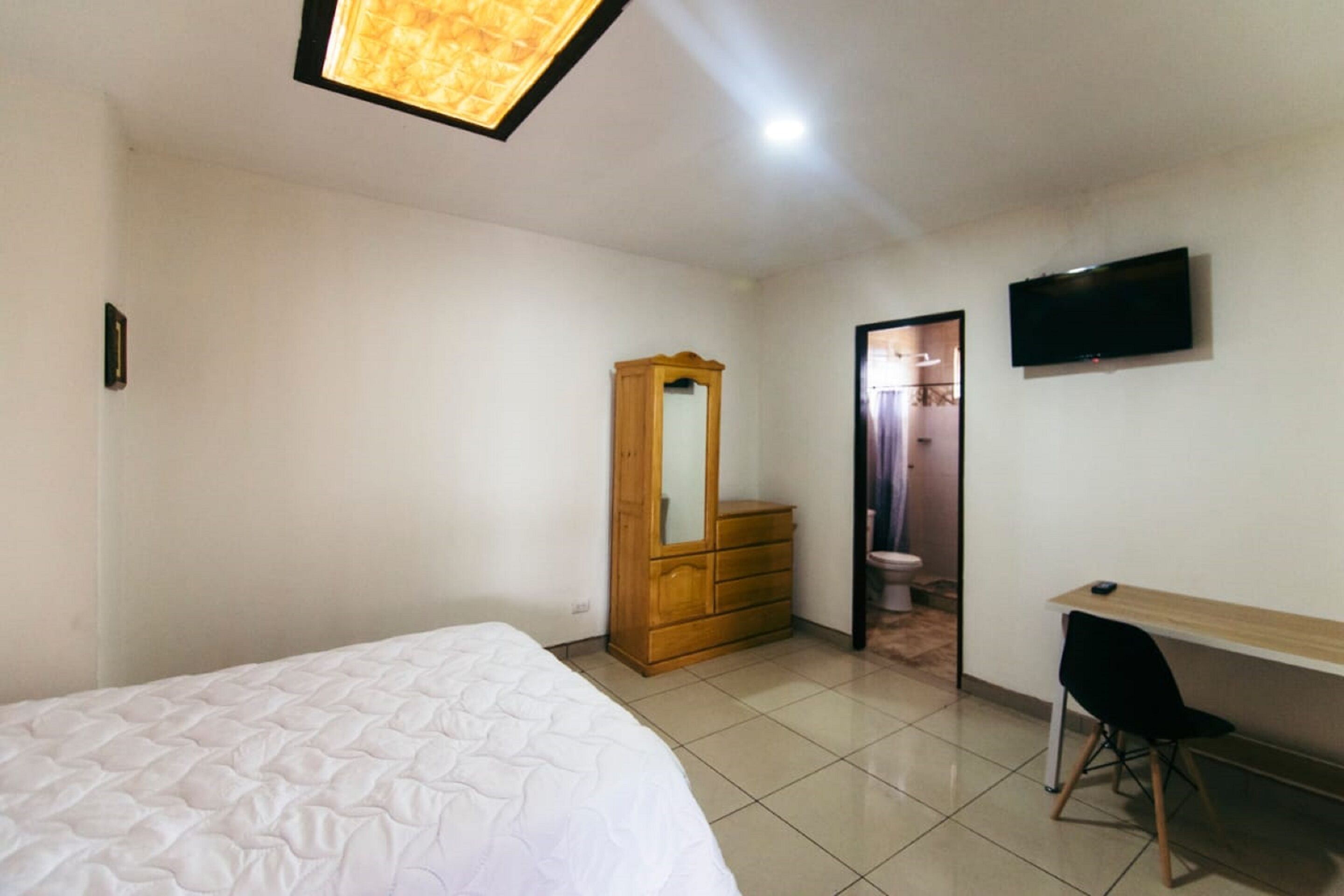 A bedroom with a bed, a wooden wardrobe, a desk with a chair, and a TV mounted on the wall.