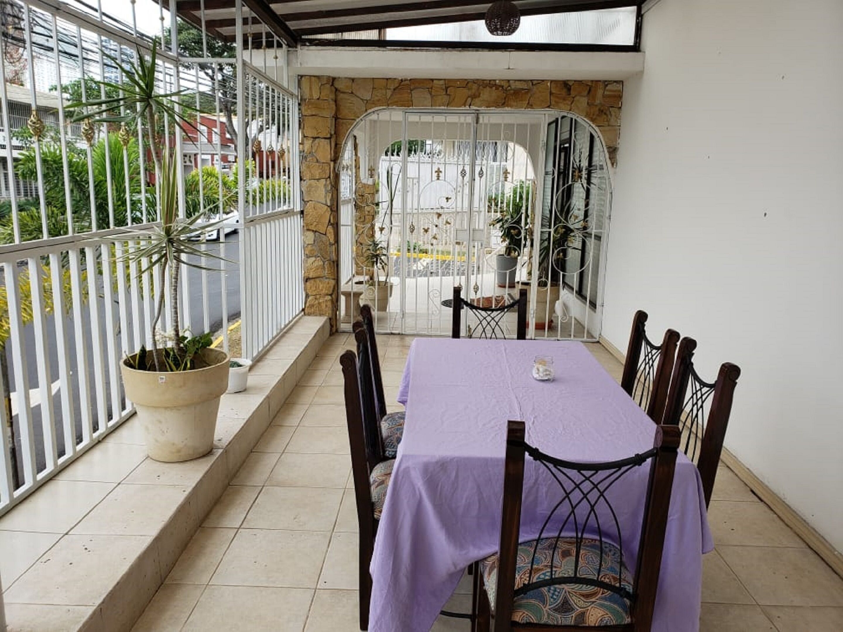 Een overdekt terras met een tafel en stoelen, een stenen muur en uitzicht op de straat.