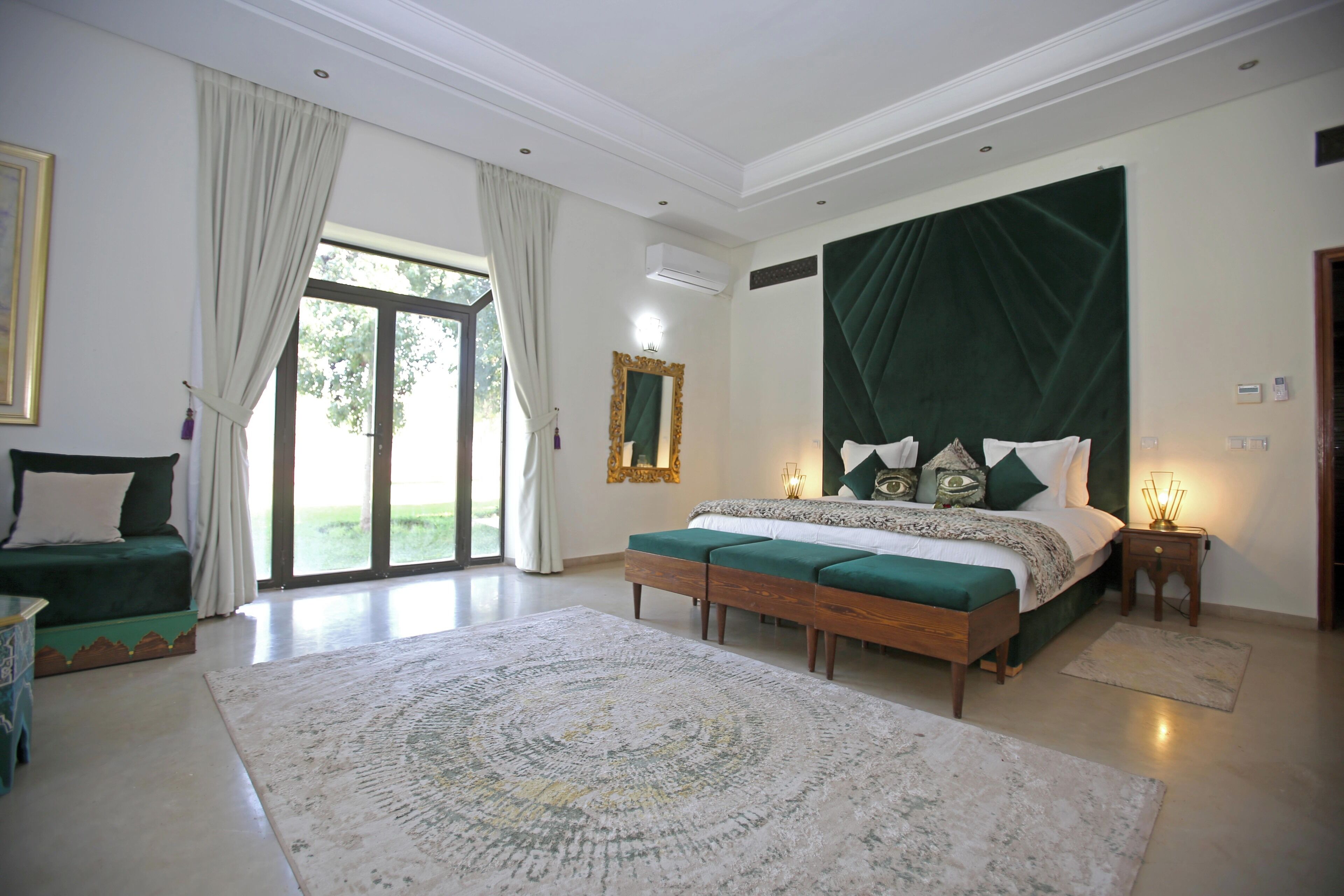 A bedroom with a large bed, a green sofa, a patterned rug, and a view through a glass door.