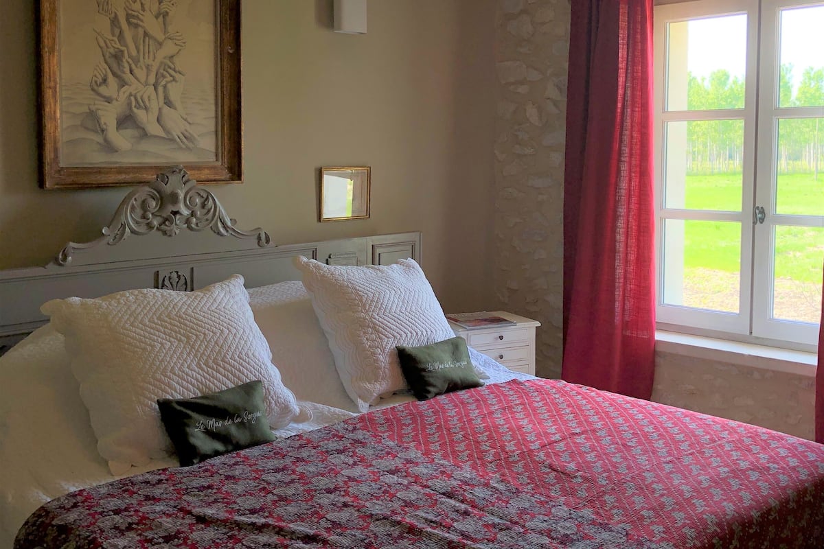 A bedroom with a bed, a nightstand, a painting, a mirror, and a window with curtains.