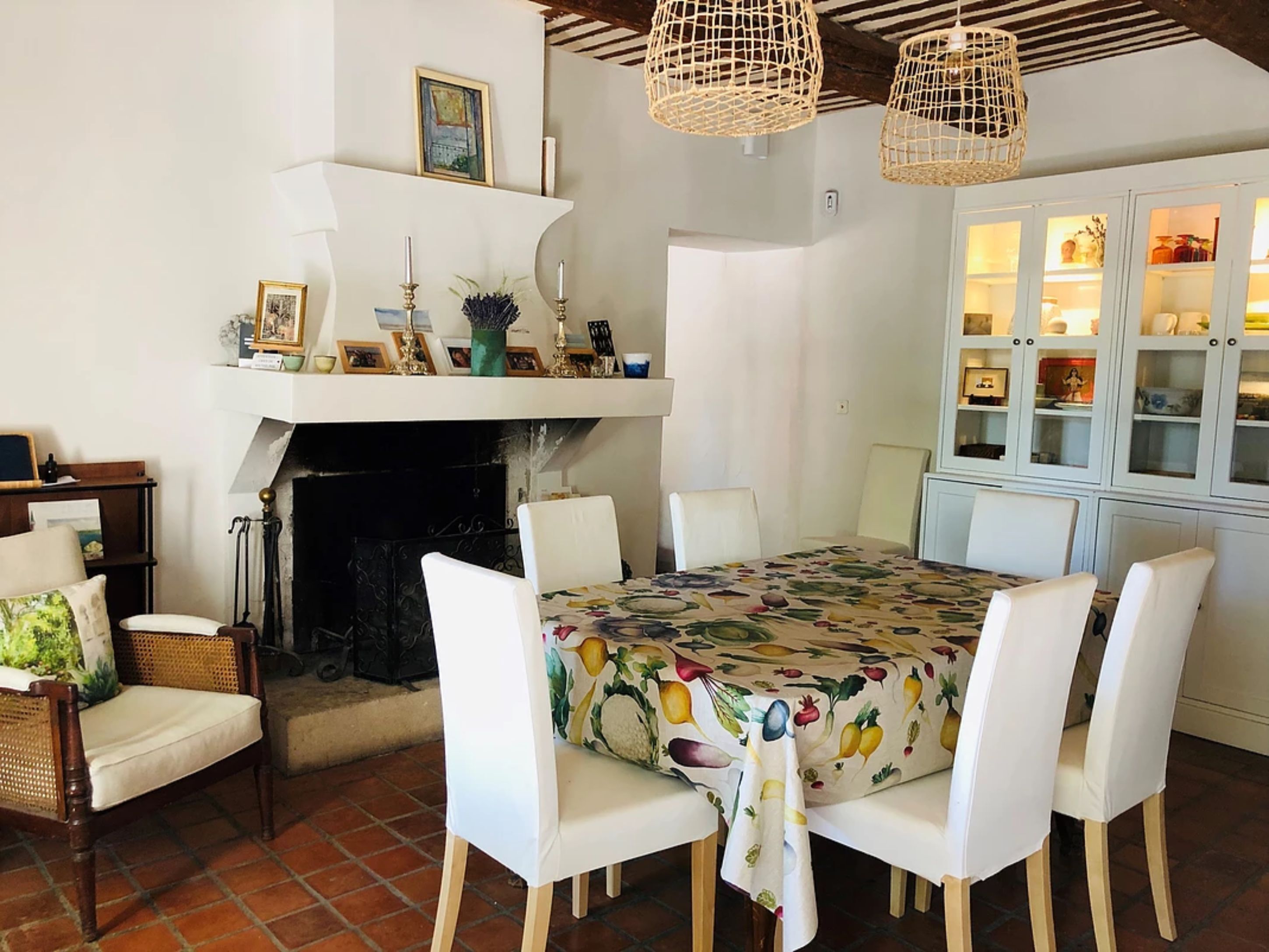 A dining area with a table covered in a fruit-patterned tablecloth, surrounded by white chairs. A fireplace is visible in the background, and there are wicker lamps hanging above the table.