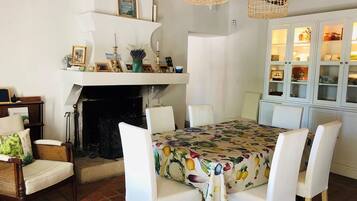 A dining area with a table covered in a fruit-patterned tablecloth, surrounded by white chairs. A fireplace is visible in the background, and there are wicker lamps hanging above the table.