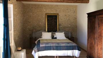 Une chambre avec un mur en pierre, un lit recouvert d’une couverture à carreaux, une armoire en bois et une petite table de chevet sur laquelle repose un chapeau.
