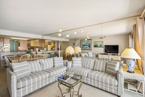 A living room with a sofa, a glass coffee table, a lamp, a TV, and a kitchen area in the background.
