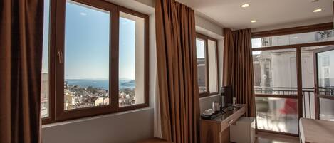 A hotel room with a bed, a desk, a chair, and a view of the city through large windows.