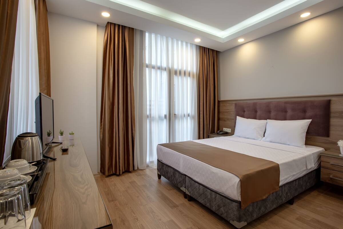 A modern hotel room with a large bed, bedside tables, a desk with a kettle, and a TV.