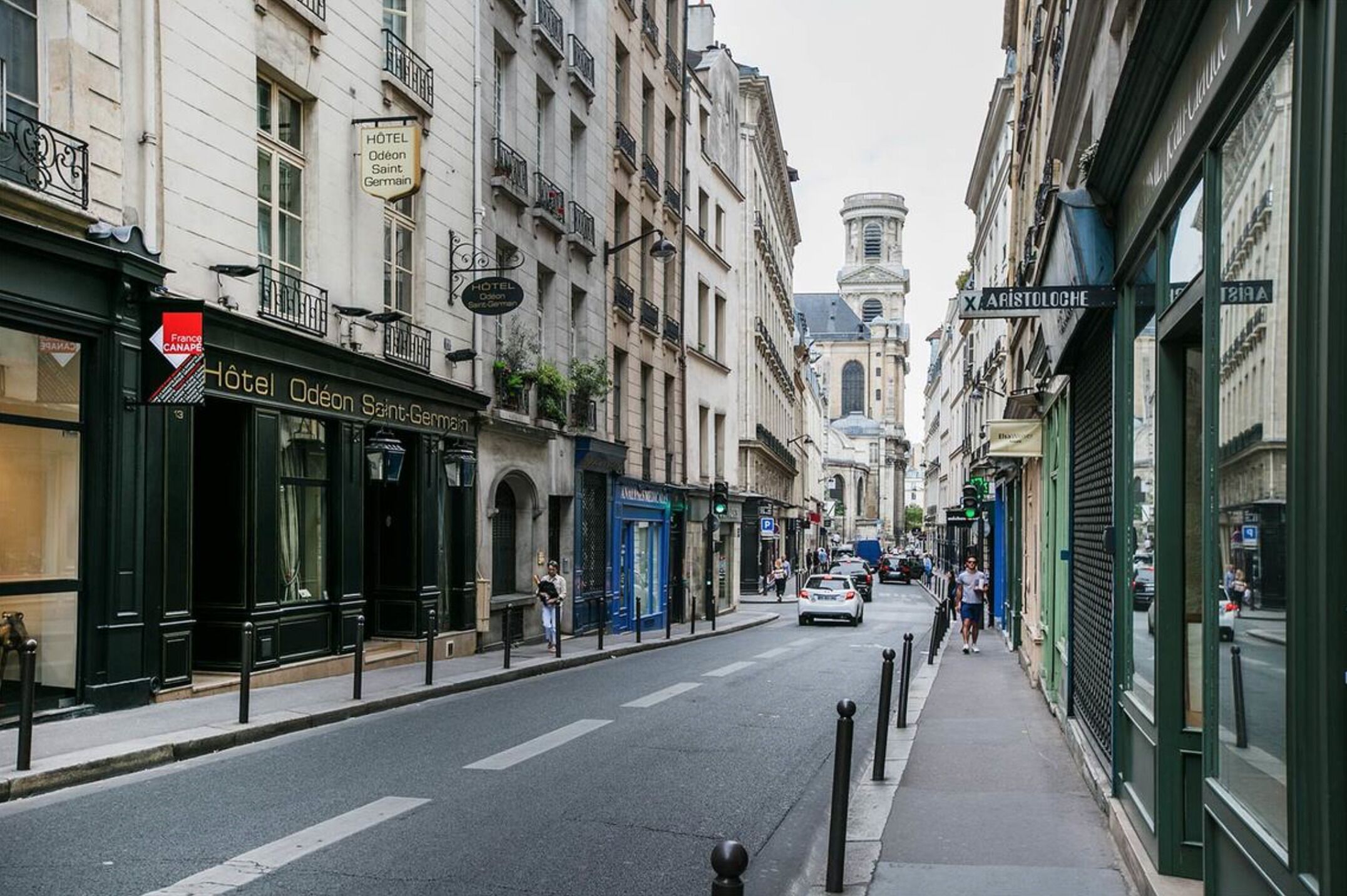 A street view with a hotel named 'Hotel Odeon Saint-Germain' on the left, featuring a black facade and a blue door.