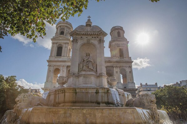 Un edificio histórico con una estatua central, dos torres de campanas y una fuente con estatuas de leones.