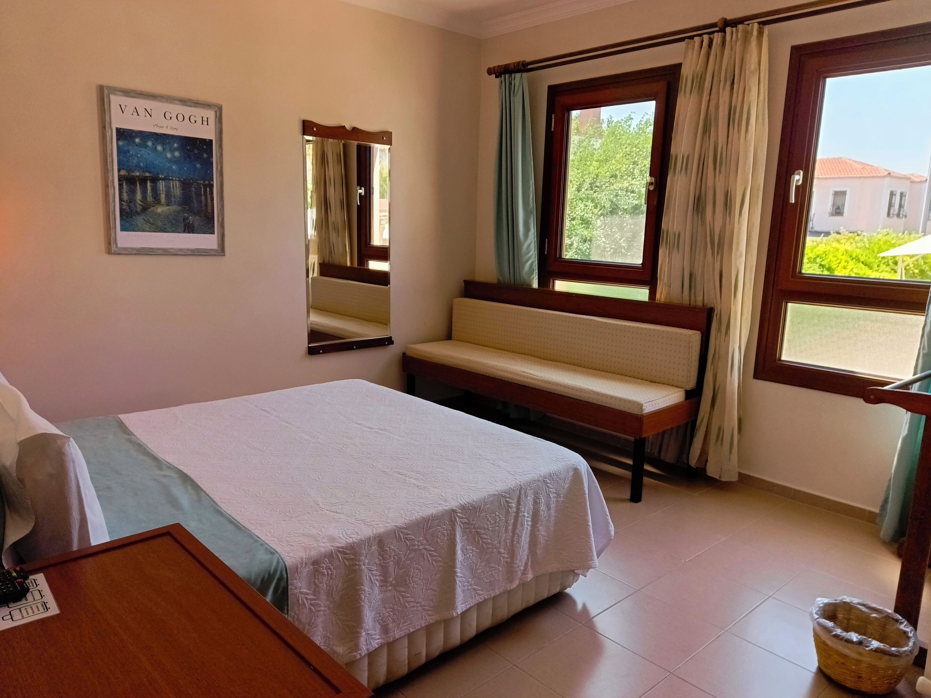 Habitación de hotel con cama, un banco, una ventana con cortinas, un espejo y un cuadro en la pared.