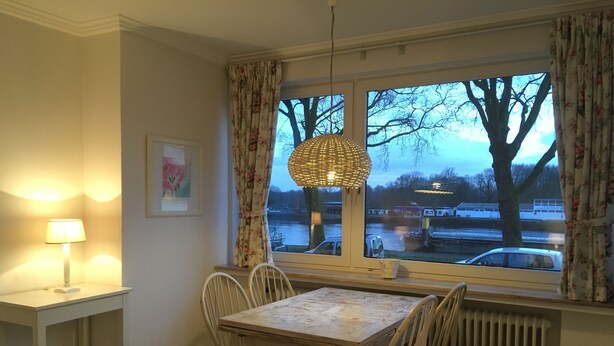 A dining area with a table and chairs, a hanging light fixture, a window with curtains, and a radiator. - Gästeträume (Bremen)