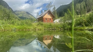 A wooden cabin with a reflection in a calm lake, surrounded by lush greenery and mountains. - Preberhütte in Prebertal lies at 1500 meters above sea level (Tamsweg)